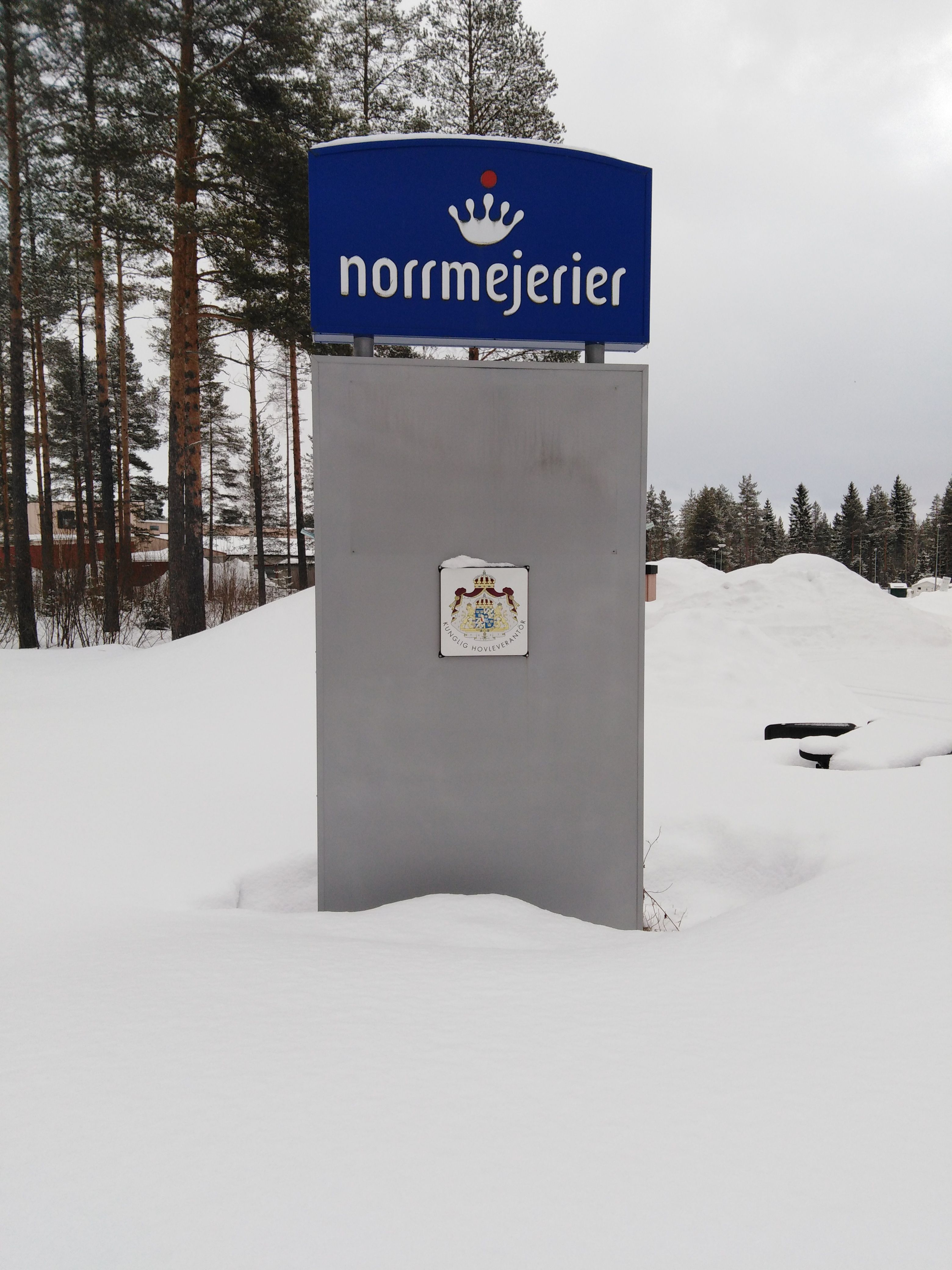 Skylt med Norrmejerier-logga omgiven av snötäckt landskap och tallar