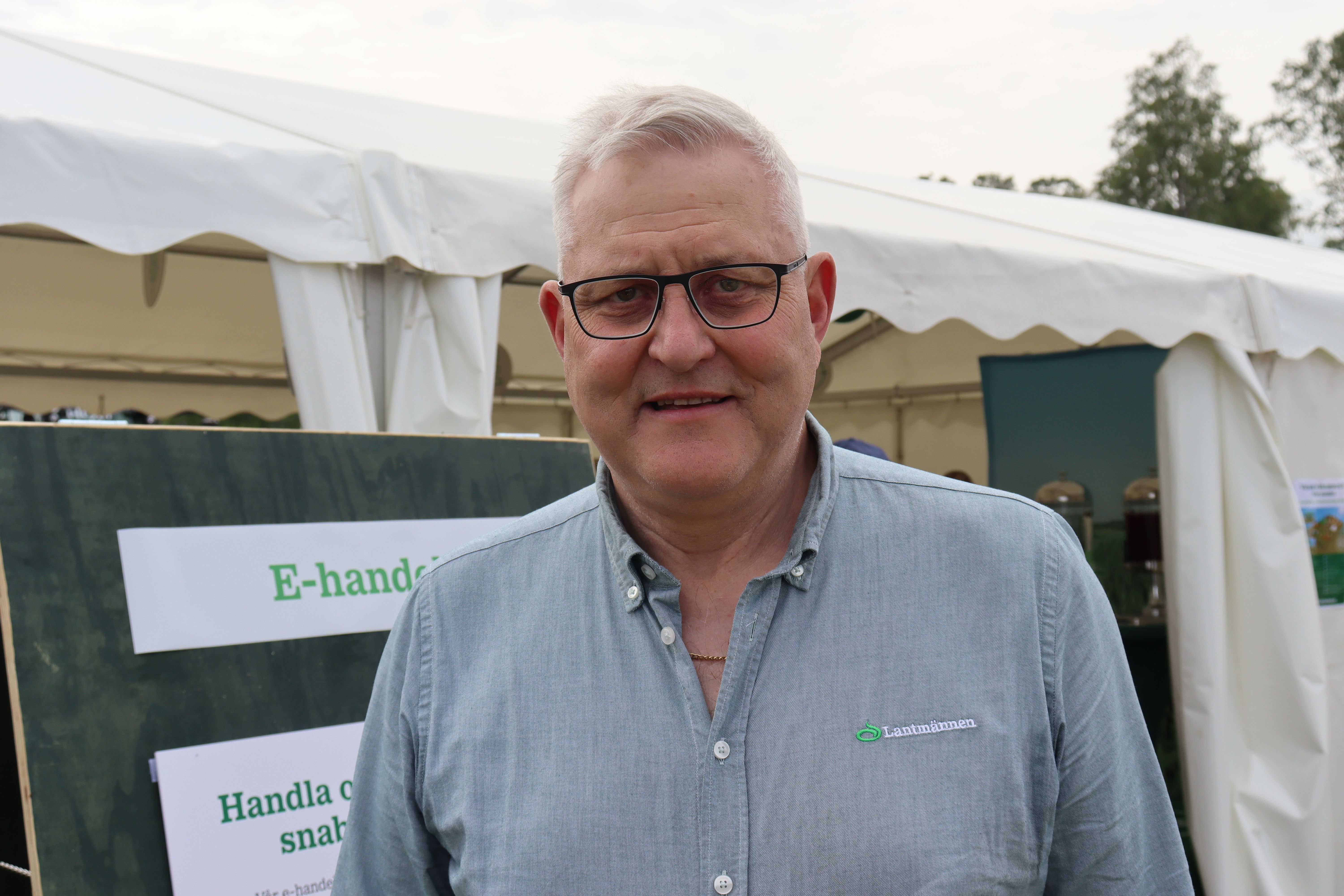 Äldre man i glasögon står framför ett vitt tält på ett utomhusevenemang