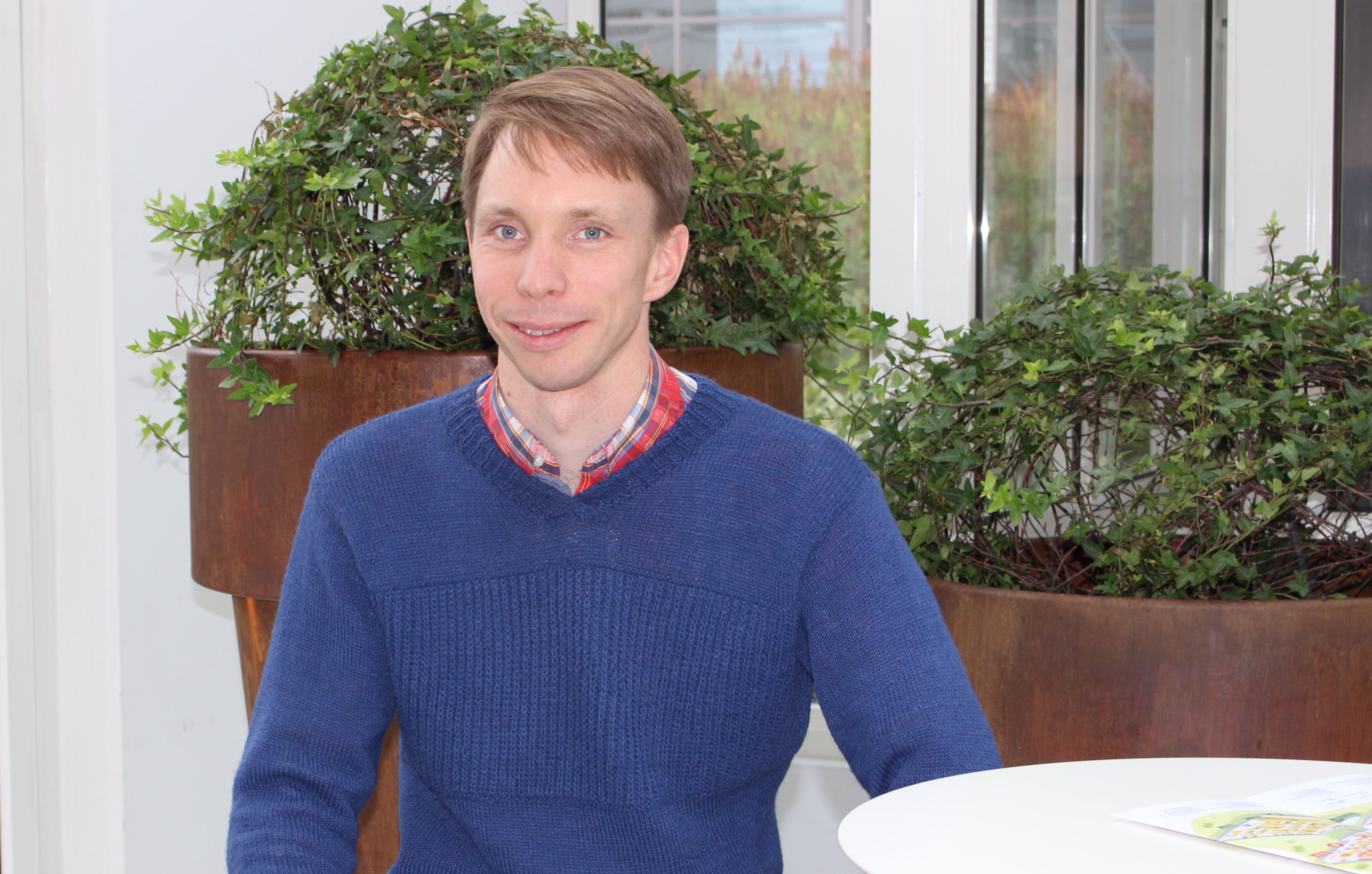 Leende man i blå tröja sitter vid vit bordsskiva framför gröna växter