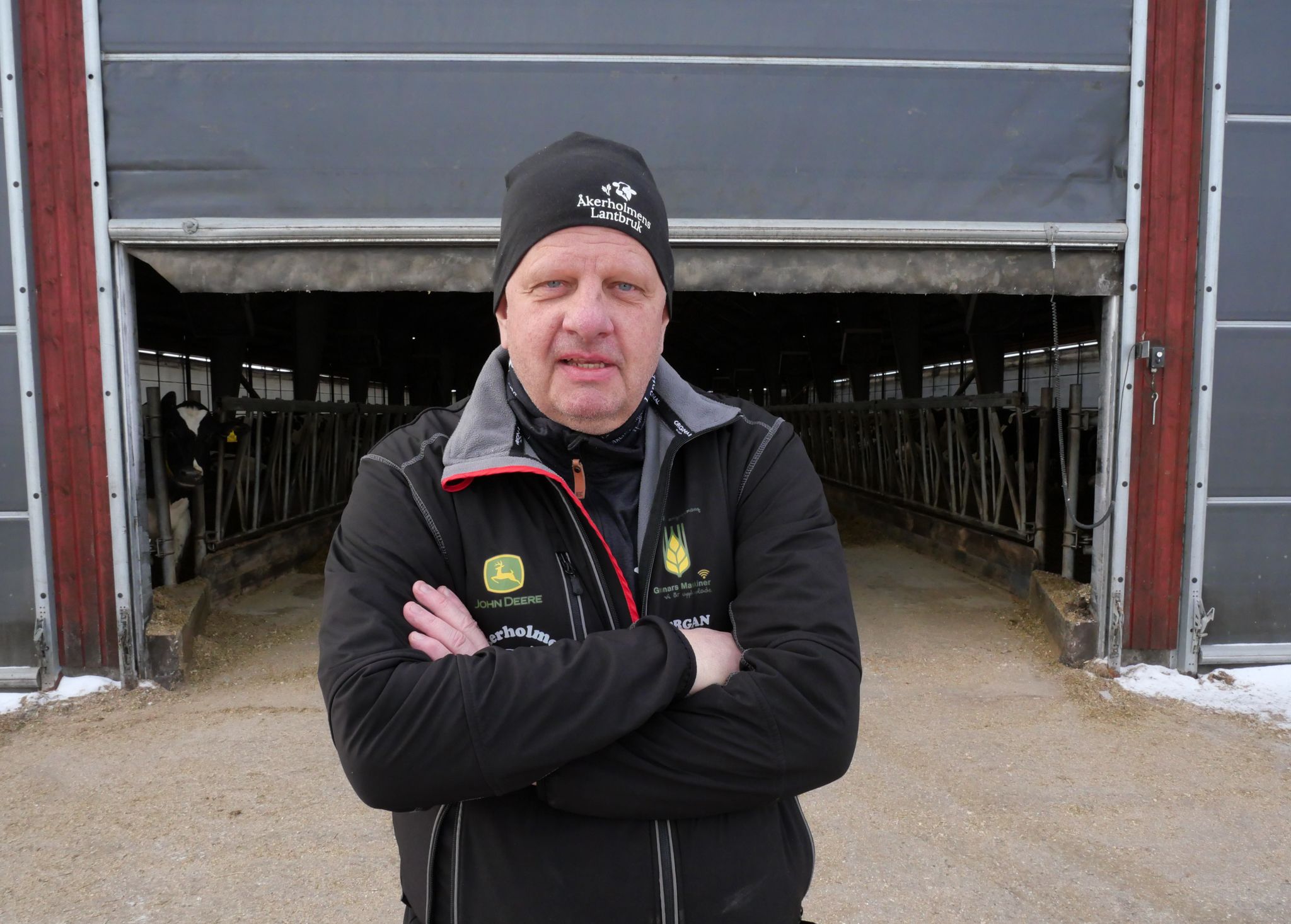 Man i arbetskläder står med korslagda armar framför en ladugård