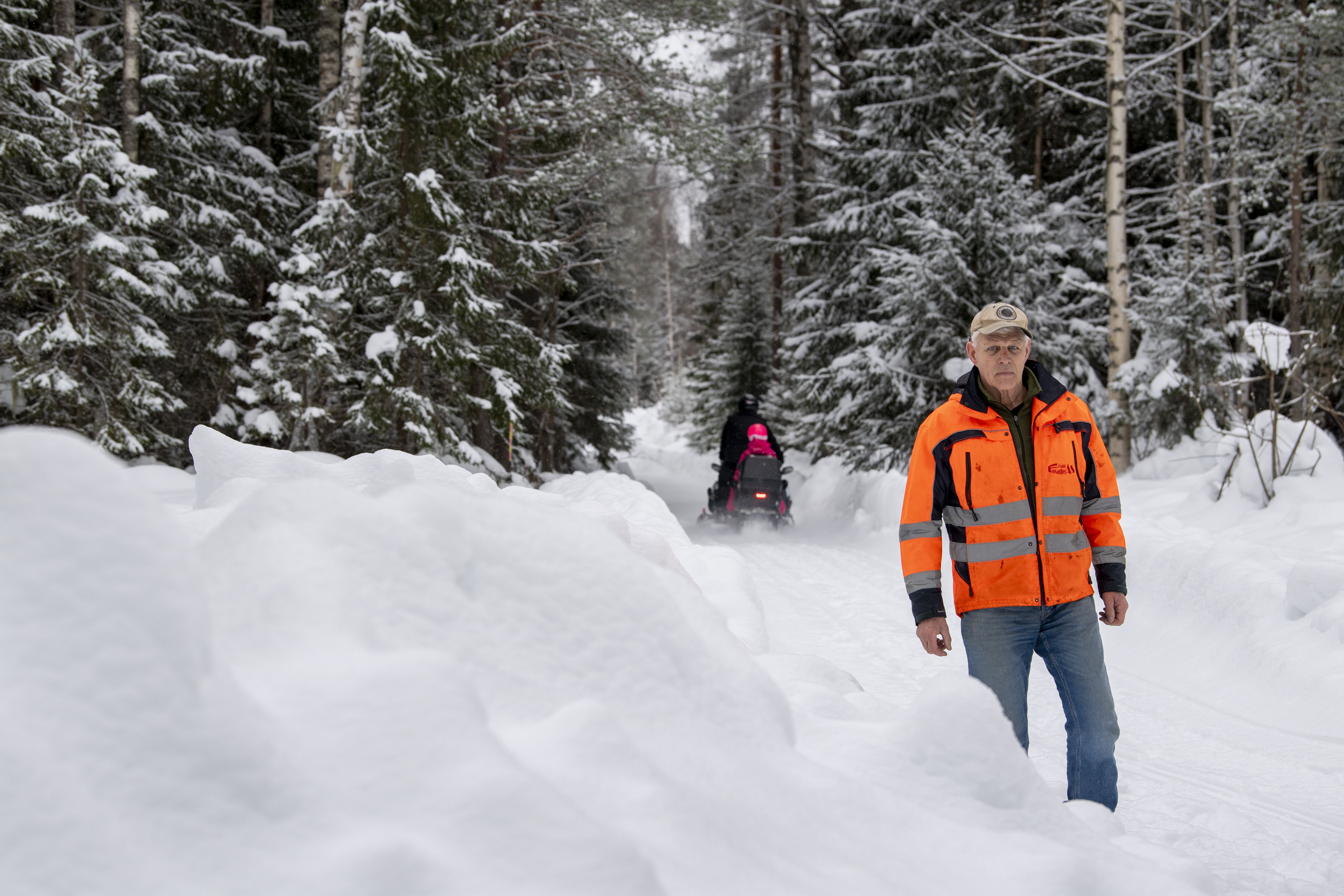 Man i orange jacka går på snötäckt skogsväg, skoter syns i bakgrunden