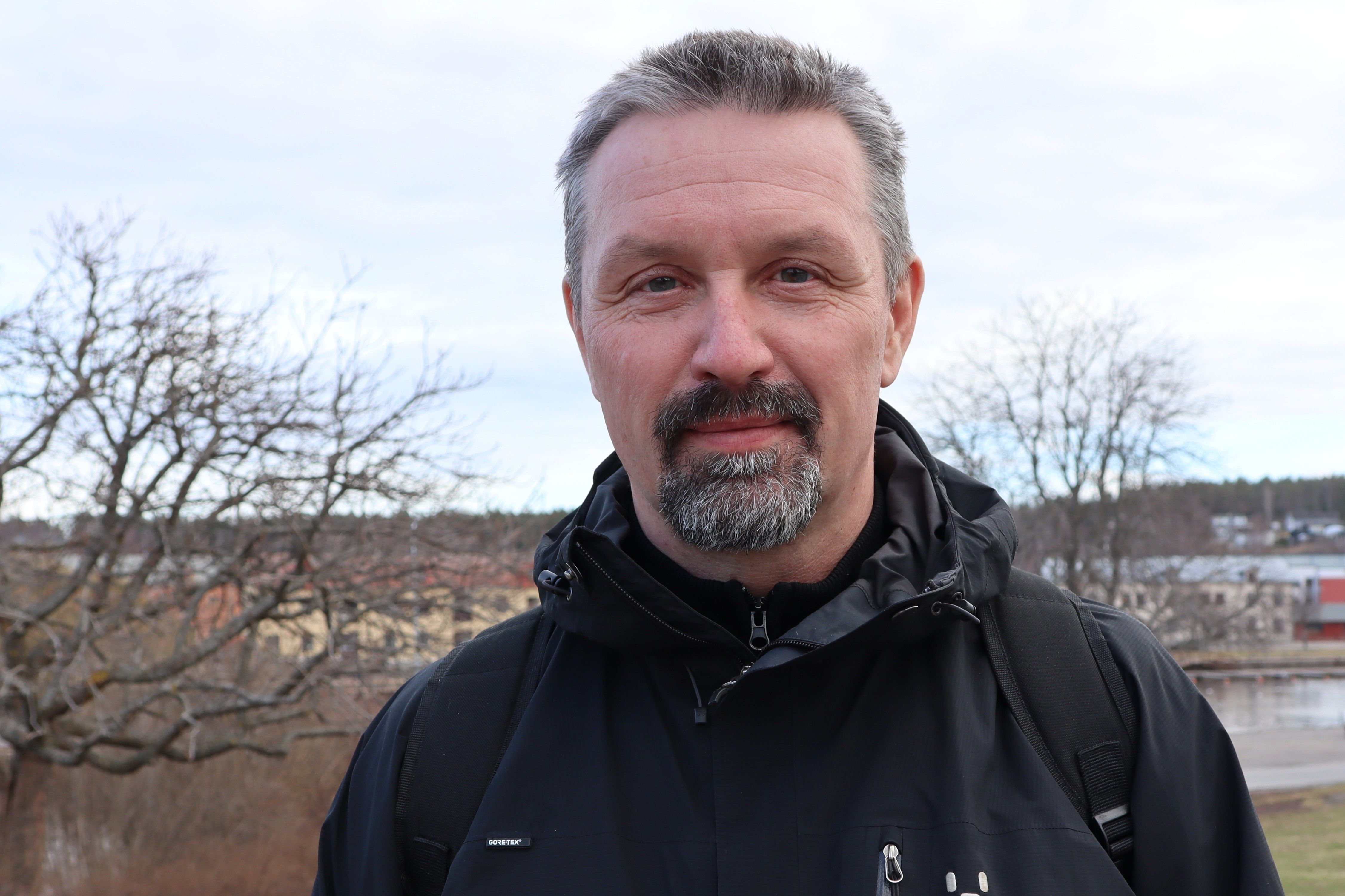 Man i svart jacka utomhus med träd och byggnader i bakgrunden