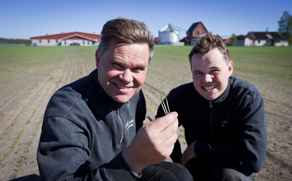 Två personer på åker håller upp små plantor framför gårdsbyggnader