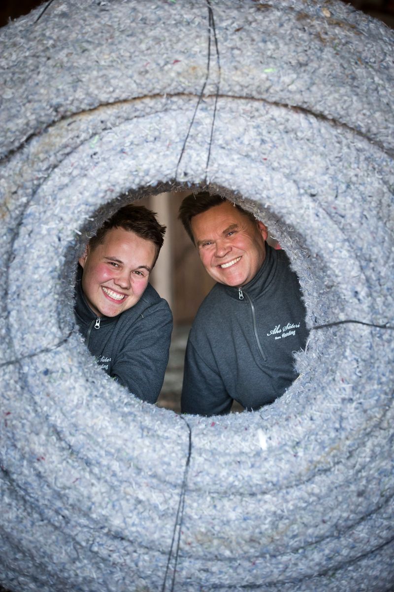 Två leende personer tittar genom ett runt hål i en stor rulle isoleringsmaterial
