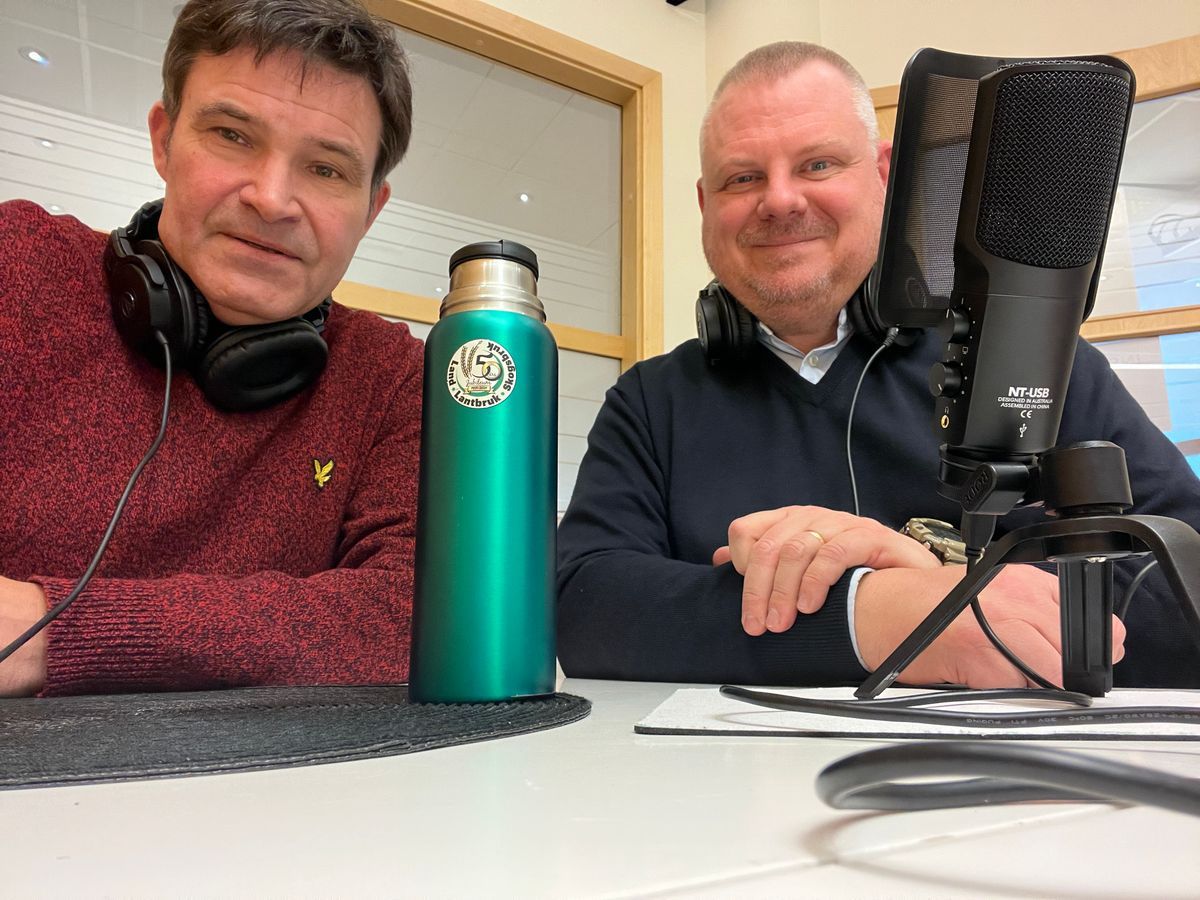 Två personer med hörlurar vid mikrofon och termosflaska i studio