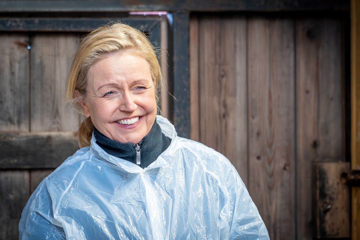 Leende kvinna i plastförkläde står utomhus framför en trävägg