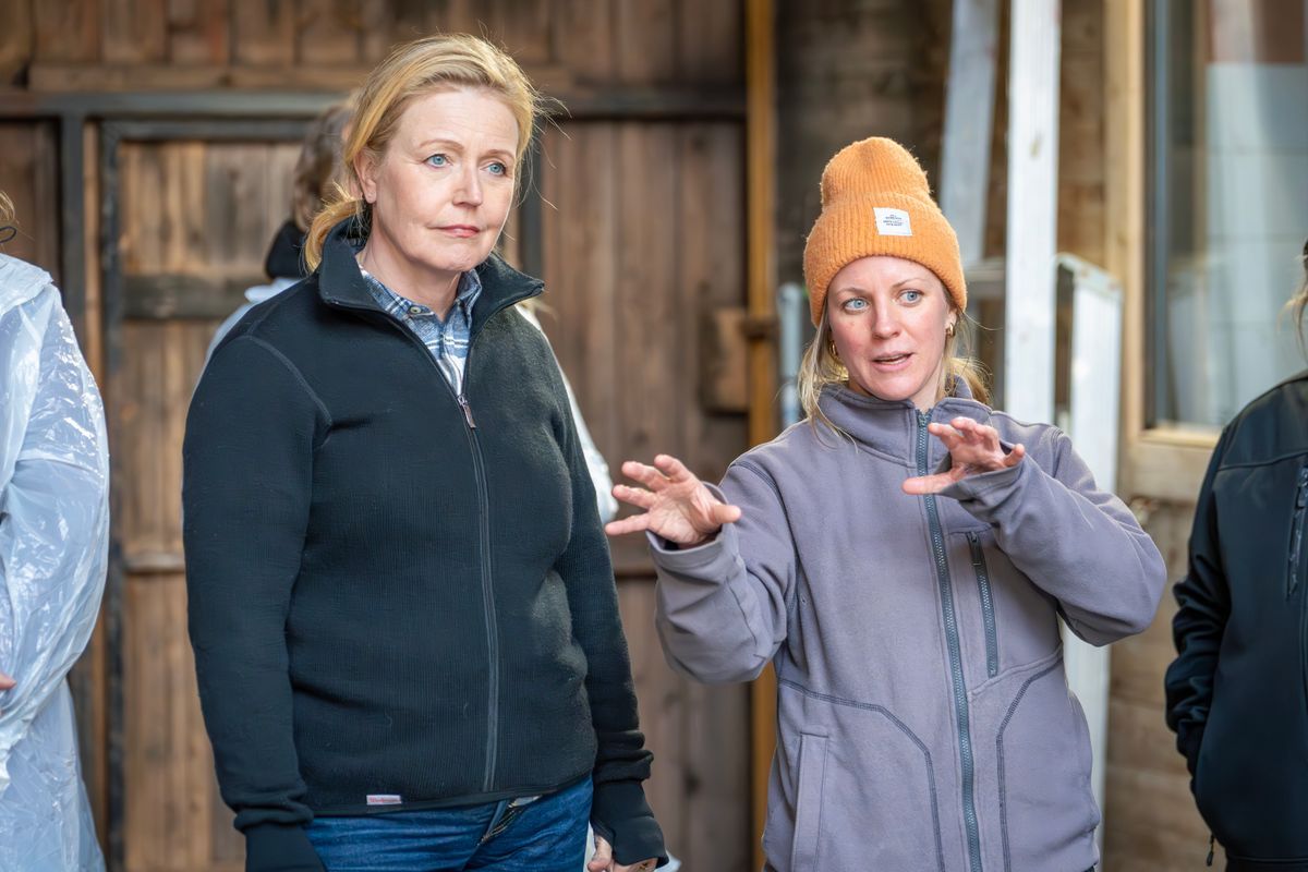 Två kvinnor i jackor samtalar, en gestikulerar med händerna i ladugårdslik miljö