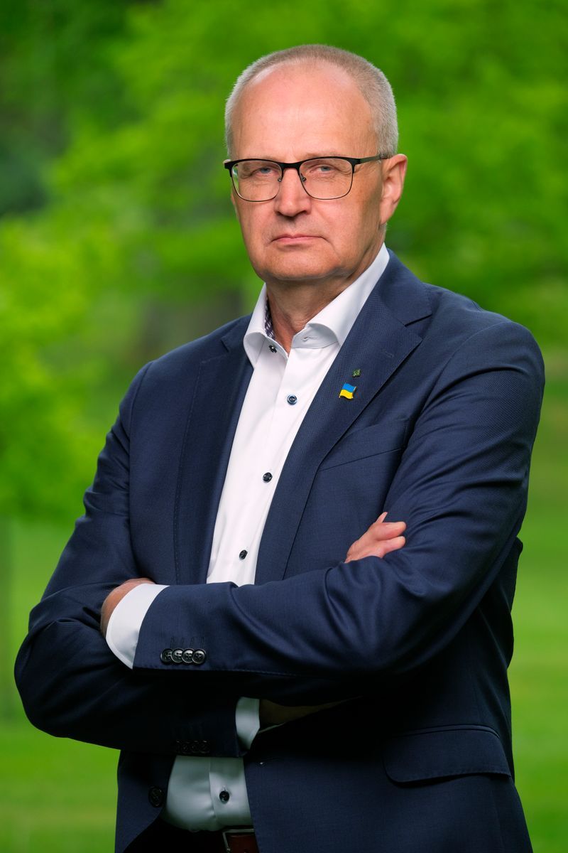 Äldre man i kostym med glasögon och korslagda armar utomhus i grön parkmiljö