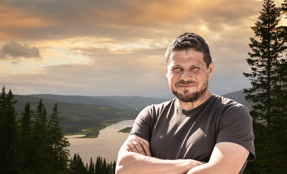 Man står med armarna i kors framför en sjö och skog i solnedgången.