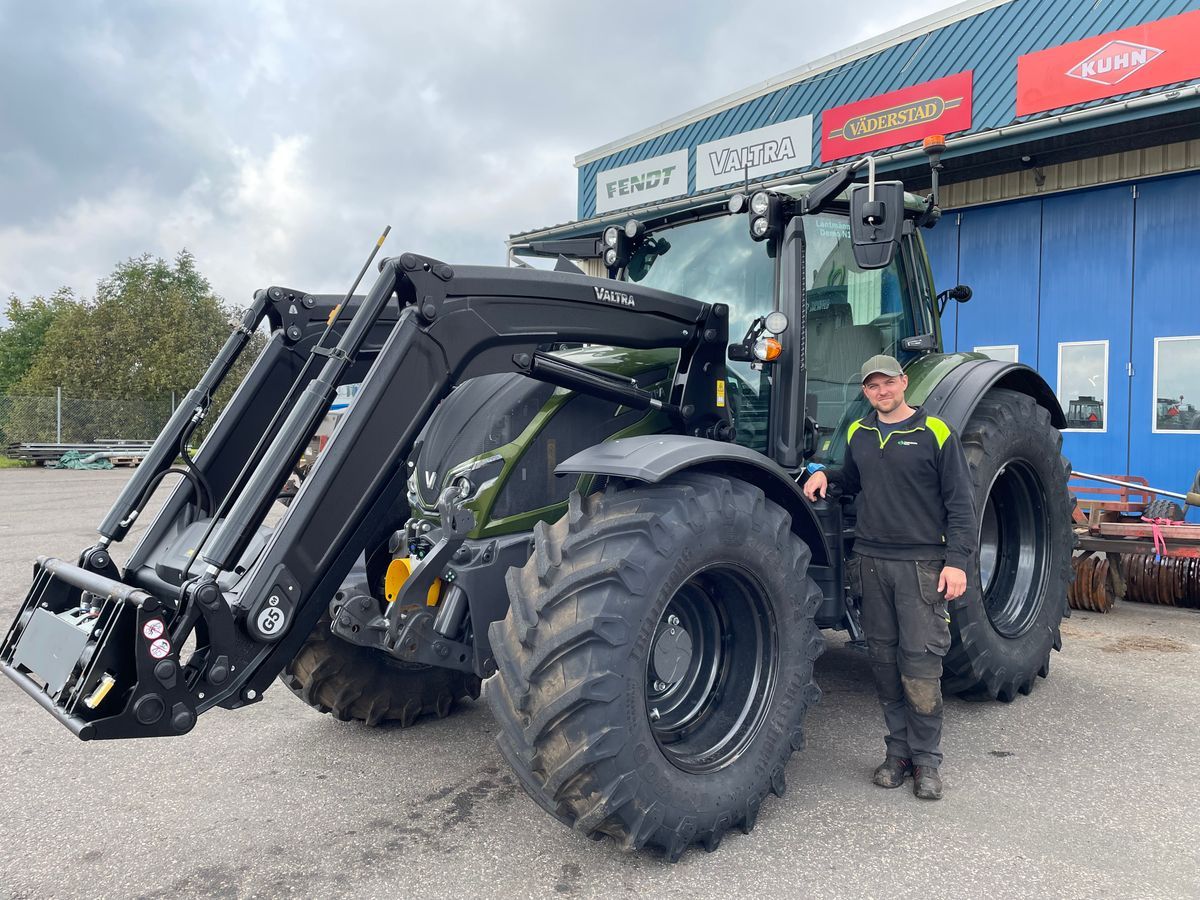 En man står bredvid en stor svart traktor med frontlastare på en gårdsplan.