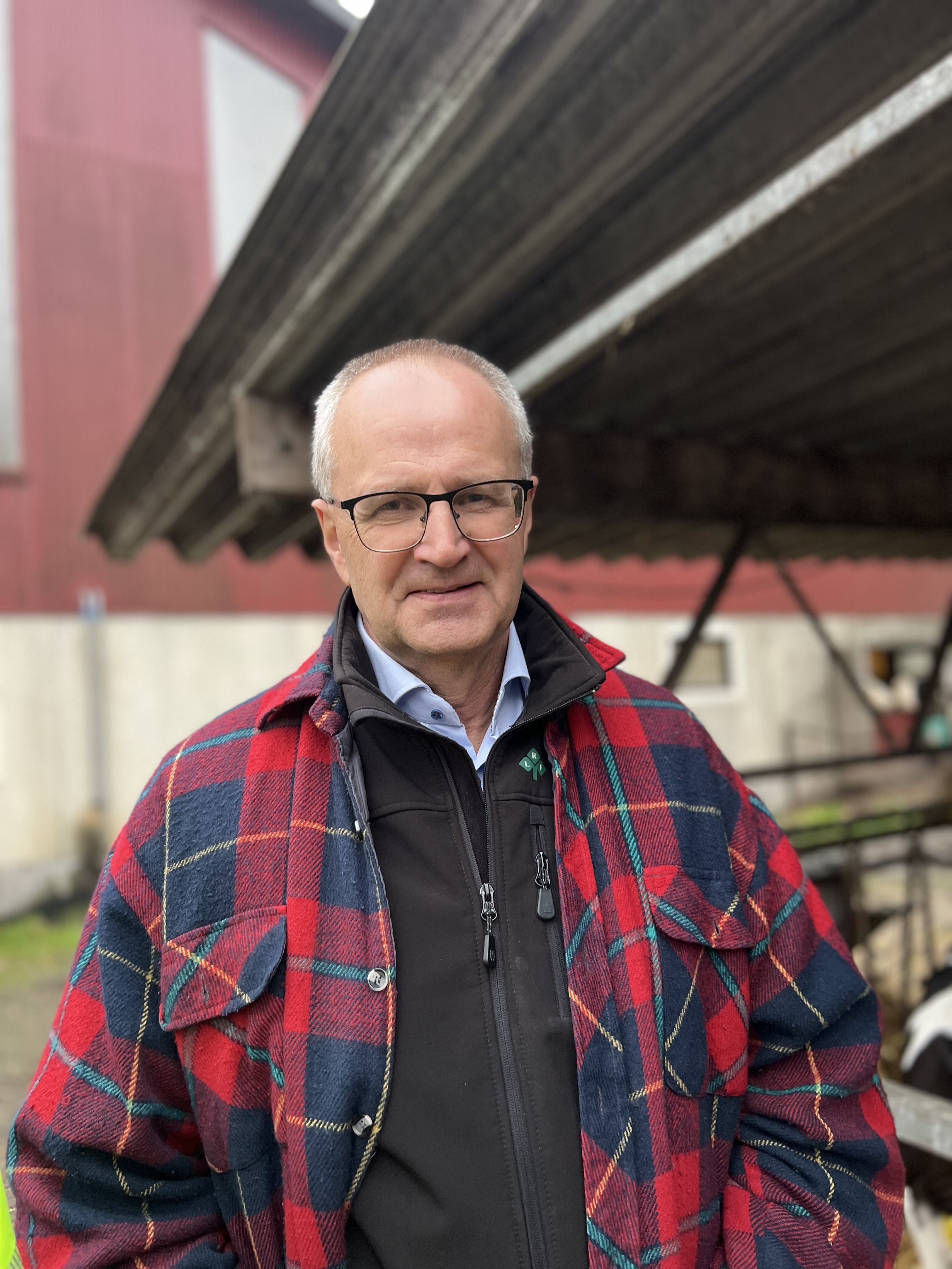 Äldre man med glasögon i rutig jacka står på en gård vid en ladugård