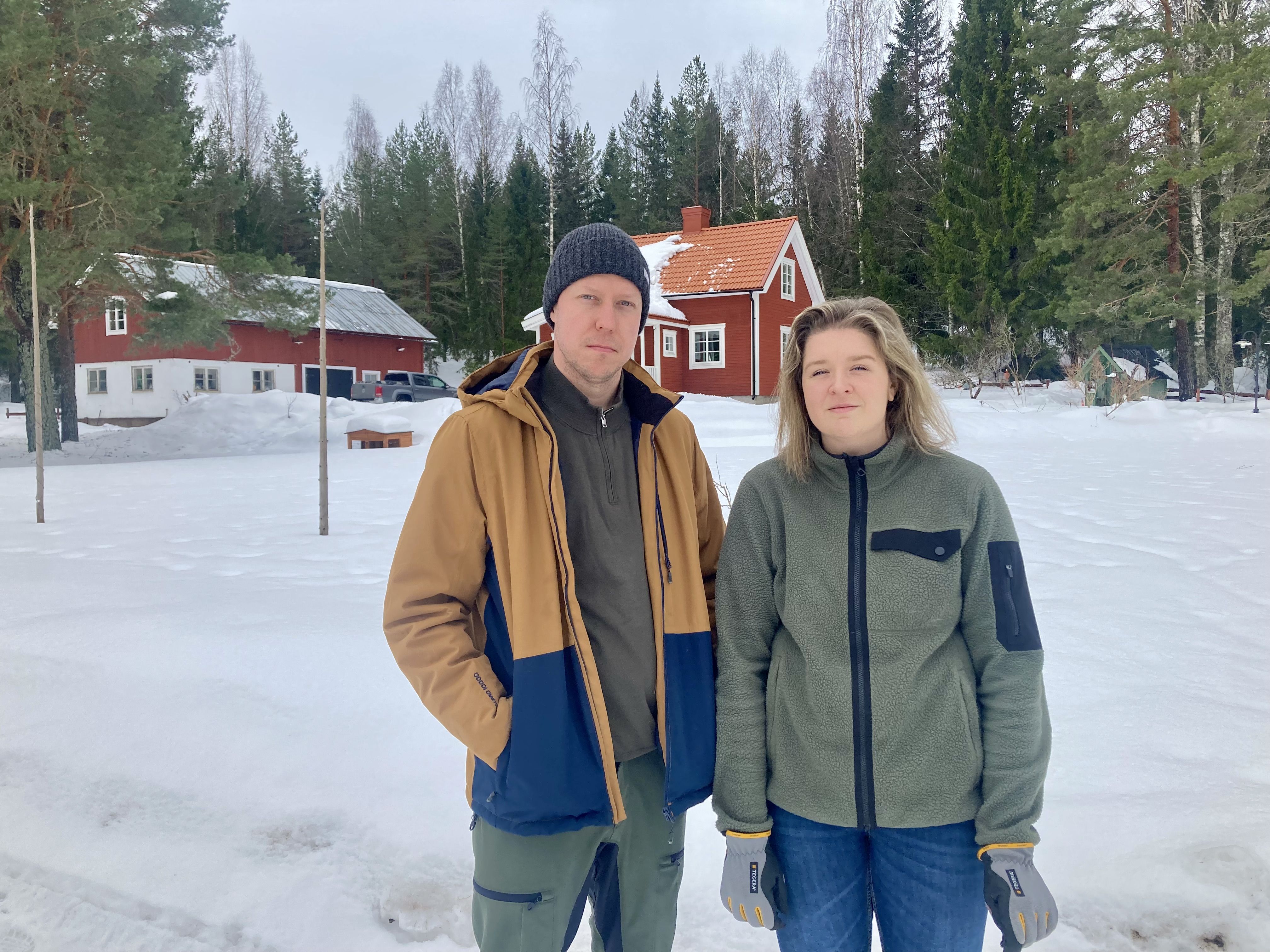 Två personer står i snötäckt trädgård framför röda hus och skog