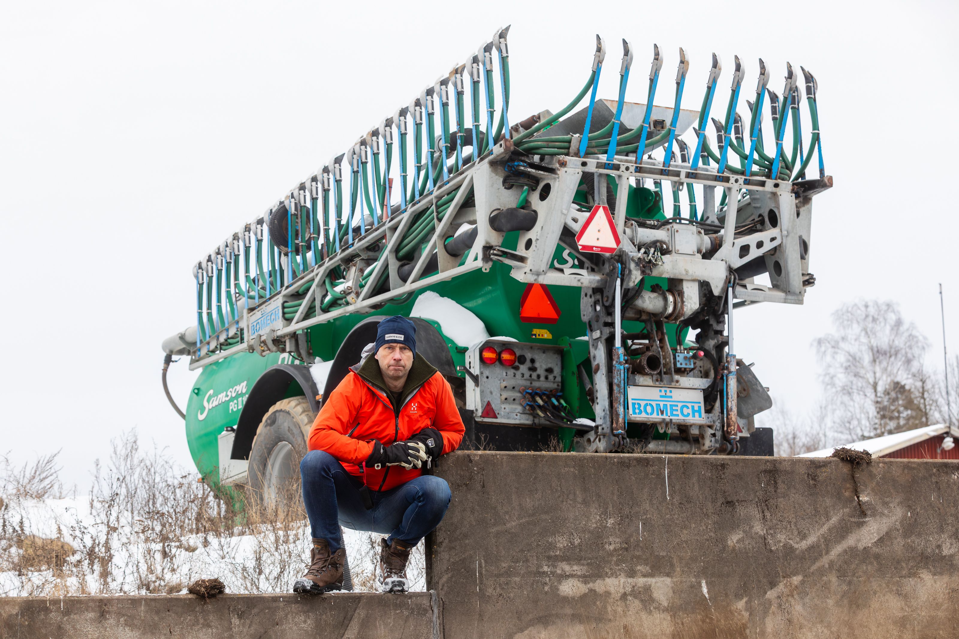 Person i orange jacka vid stor grön gödselspridare en vinterdag